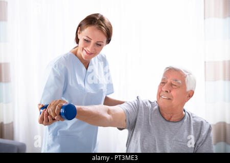 Young Female Nurse Helping Happy Senior Man With Dumbbell Exercise Stock Photo