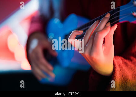 Hands of young pretty girl playing on blue ukulele while sitting on glowing neon stairs in amusement park at night Stock Photo