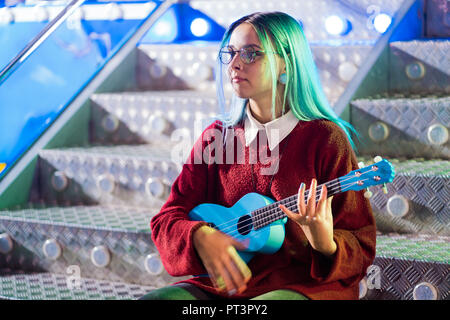 Young pretty girl with blue hair playing on blue ukulele while sitting on glowing neon stairs in amusement park at night Stock Photo