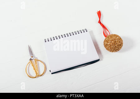 Stylish flat lay new year's arrangement of preparation for holiday. Top view blank open notebook near golden christmas ball and scissors on white wood Stock Photo