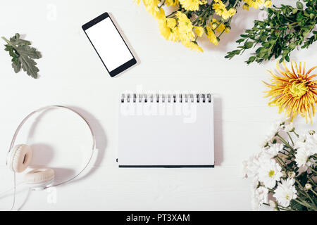 Floral flat lay composition with smart device, earphones and empty notebook in center. Top view of open blank notepad sheet near mobile phone, headpho Stock Photo