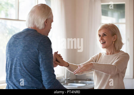 Old happy couple dancing in kitchen, mature family celebrating anniversary having fun together at home, middle aged caring husband holding hands of ex Stock Photo