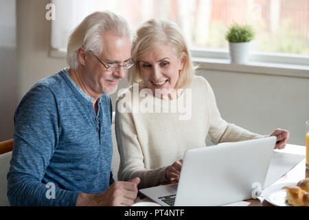 Happy old family couple talking using laptop having breakfast together, surprised excited senior woman looking at computer screen showing smiling midd Stock Photo