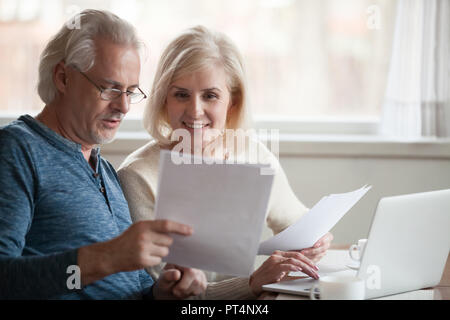 Happy old middle aged couple holding reading good news in document, smiling senior mature family excited by mail letter, checking paying domestic bill Stock Photo