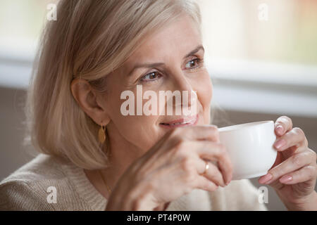 Thoughtful happy middle aged senior woman with beautiful face looking away drinking morning coffee, smiling mature old lady holding tea cup relaxing w Stock Photo