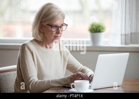Serious mature middle aged business woman in glasses using laptop typing email working at home office, thoughtful focused senior old lady searching in Stock Photo
