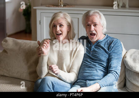 Surprised senior middle aged couple viewers feeling amazed watching new reality show on television channel, astonished shocked old mature family looki Stock Photo