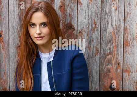 Outdoor portrait of beautiful girl or young woman with red hair wearing a ble jacket and white t-shirt leaning against an old wooden building Stock Photo