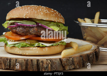 A big burger with a meat fried chop, tomatoes, pickled cucumbers, cheese, onions and salad. Next to French fries on a wooden piece of wood. Stock Photo