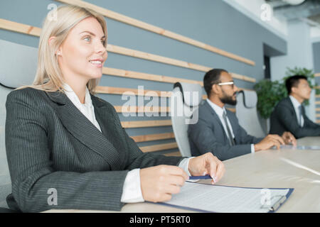 Smiling successful attractive business lady in formal jacket sitting at table with clipboard and listening to conference speaker at meeting Stock Photo