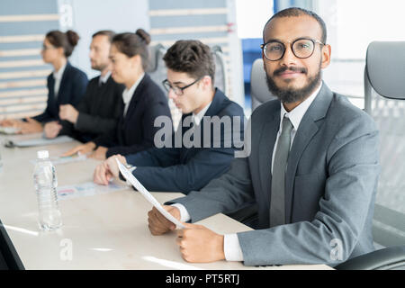 Content handsome young Arabian businessman in formal suit sitting at table in line with colleagues and looking at camera while checking paper at confe Stock Photo