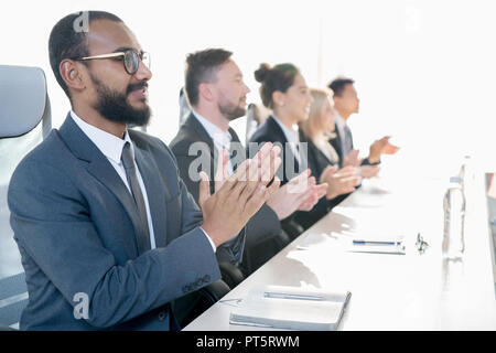 Group of positive excited business people sitting in line and applauding while greeting coach at conference Stock Photo