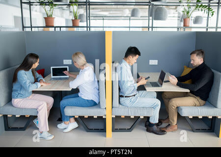 Creative colleagues discussing project in coworking space: busy ladies using laptop and analyzing data together, guys using smartphones Stock Photo