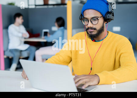 Serious concentrated young middle-eastern man with beard and mustache wearing headphones sitting at table and working with laptop Stock Photo