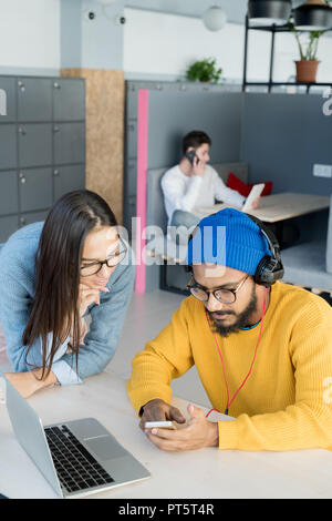 Serious concentrated hipster middle-eastern man in headphones sitting at table and showing new mobile app to colleague in modern office Stock Photo