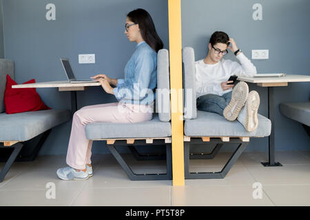Serious working lady in glasses using modern laptop while pensive young guy relaxing on sofa and using smartphone in cafe Stock Photo