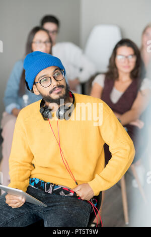 Serious pensive young Arabian student with beard wearing bright hat and sweater sitting on chair and holding tablet for notes while listening to speak Stock Photo