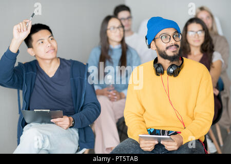 Group of content curious multi-ethnic students participating in discussion at master class and sitting on chairs, Asian guy raising hand to ask questi Stock Photo