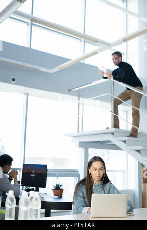 People working in modern office: serious woman listening to music and using laptop at table, busy man leaning on railings and analyzing information on Stock Photo