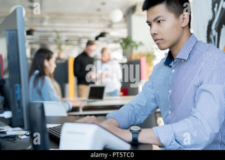 Serious busy young Asian manager in formal shirt sitting at desk and typing on computer keyboard while taking statistics Stock Photo