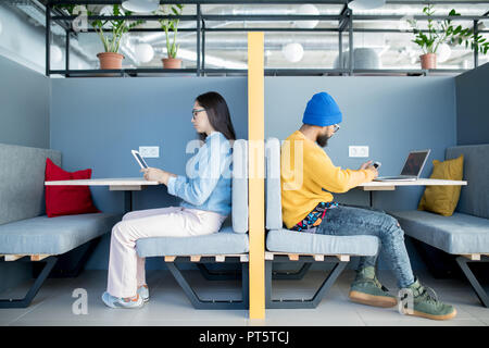 Serious concentrated hipster young people sitting at tables beyond wall and using modern devices while working in open space Stock Photo