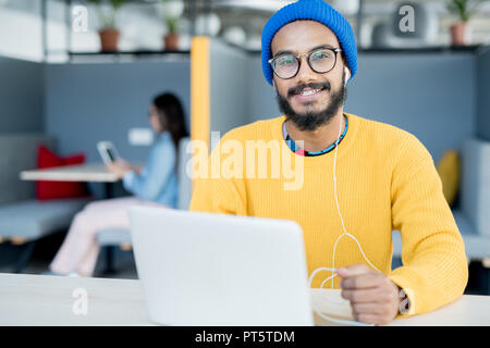Happy excited young Arabian guy in earphones wearing blue knitted hat and yellow sweater sitting at table and using laptop while looking at camera in  Stock Photo