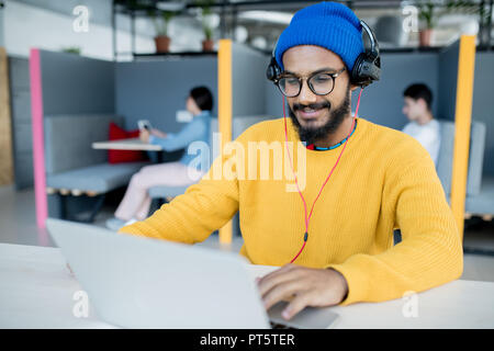 Positive satisfied middle-eastern app developer in headphones sitting at table and using laptop while working in open space office Stock Photo