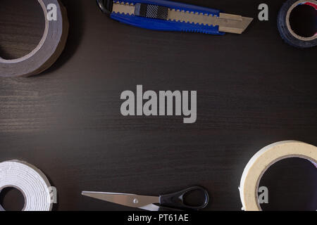 A black wooden background with various cutting tools: scissors, utility knife, tape. Stock Photo