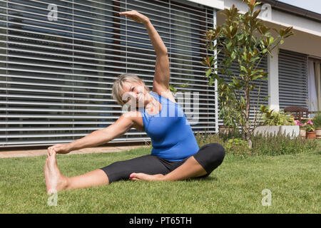 Flexible attractive senior blond woman is fitness personal instructor exercising sitting on the lawn and smiling at the camera. Stock Photo