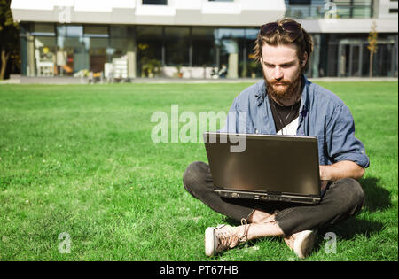 Handsome bearded student in jeans shirt wearing sunglasses sitting on the green grass near the university and doing his homework with laptop Stock Photo