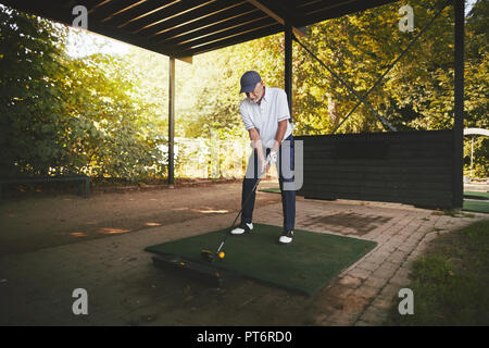 Sporty senior man standing at a tee practicing his golf swing at a driving range on a sunny day Stock Photo