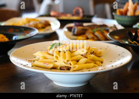 Food concept. Pasta penne with mushroom, bacon and whipping cream on white plate. Blur plates with foods background. Stock Photo