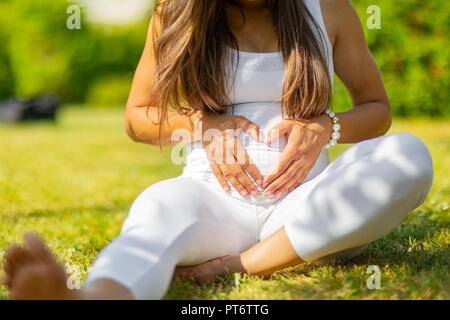 Close-up of pregnant woman sitting outdoor making hand heart gesture on belly Stock Photo