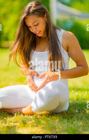 Beautiful pregnant woman sitting outdoor making hand heart gesture on belly Stock Photo