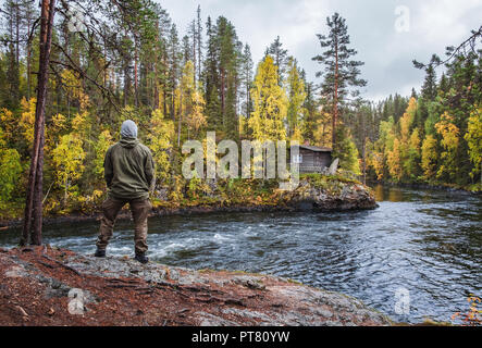 Man standing front of beautiful forest with fall colors and old cottage at mood day in Myllykoski, Kuusamo, Finland Stock Photo
