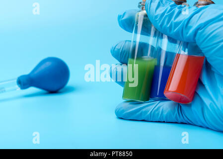 A gloved hand holds three vials of different liquid inside on a blue background. Tubes with red, green and blue liquid. Stock Photo