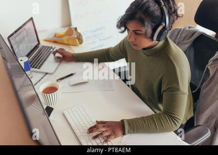 Female programmer coding on a desktop computer and laptop at her desk in office. Young woman in casuals wearing headphones working on a laptop and des Stock Photo