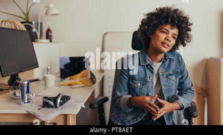 African woman sitting at her desk and listening to colleague at tech startup. Female computer programmer discussing work with team. Stock Photo