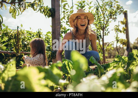 Beautiful young female farmer working in her garden with her daughter sitting by. Young mother and daughter working in the farm. Stock Photo
