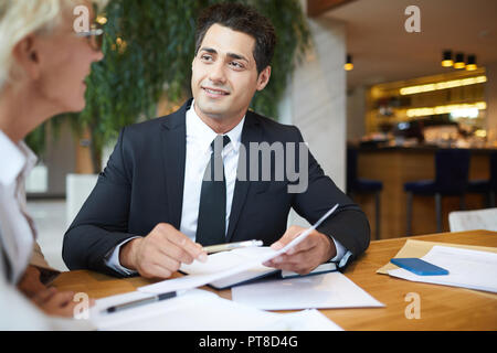 Cheerful confident handsome young businessman in formal suit meeting with business partner in restaurant: he explaining contract conditions and showin Stock Photo