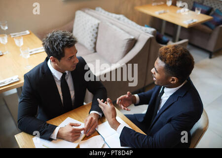Serious confident multi-ethnic businessmen in suits sitting at table and discussing strategies in cafe: African man gesturing hands while explaining p Stock Photo