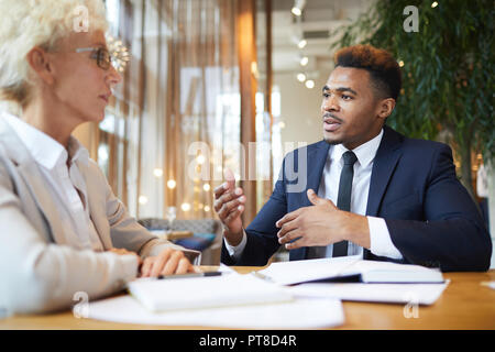 Serious confident multi-ethnic business colleagues in formalwear sitting at table and brainstorming about new project, black man sharing ideas with la Stock Photo