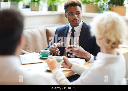 Confident successful black manager in suit sitting at table with coffee cups and laptop and gesturing hands while sharing development strategy with co Stock Photo