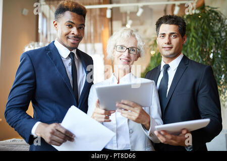 Cheerful confident female head of company and her team using modern device and examining paper together, they looking at camera Stock Photo