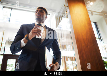 Below view of serious confident modern businessman in formal suit holding laptop and drinking coffee from disposable cup while standing in lobby Stock Photo