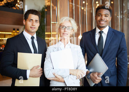 Professional business team: smiling attractive mature female head of company and her colleagues holding papers and laptop while looking at camera in l Stock Photo