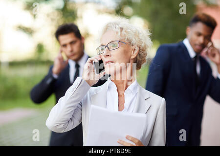 Serious confident mature lady lawyer with curly hair wearing eyeglasses calling client on mobile phone while viewing papers, businessmen talking by ph Stock Photo