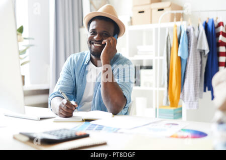 Young cheerful African-american man in smart casual looking at you while consulting someone on smartphone by workplace Stock Photo