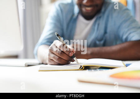 African-american creative guy with pen making working notes in notebook while sitting by desk Stock Photo