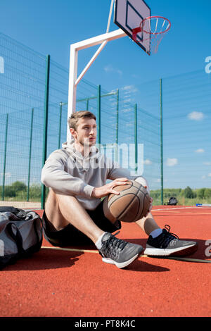 Full length portrait of contemporary young  man sitting on floor at basketball court holding ball, copy space Stock Photo